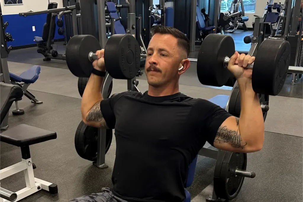 A man with a mustache, working out with 2 heavy dumbbells.