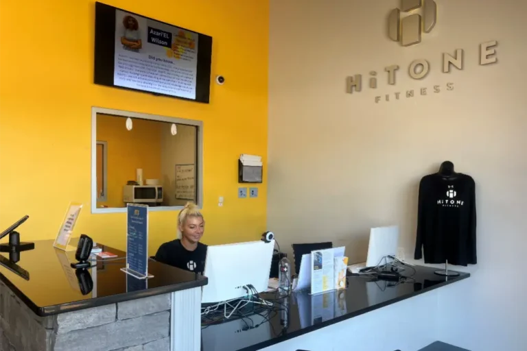 A blonde woman sitting at a reception desk at the HiTONE fitness gym. There are a mirror and a screen behind her.