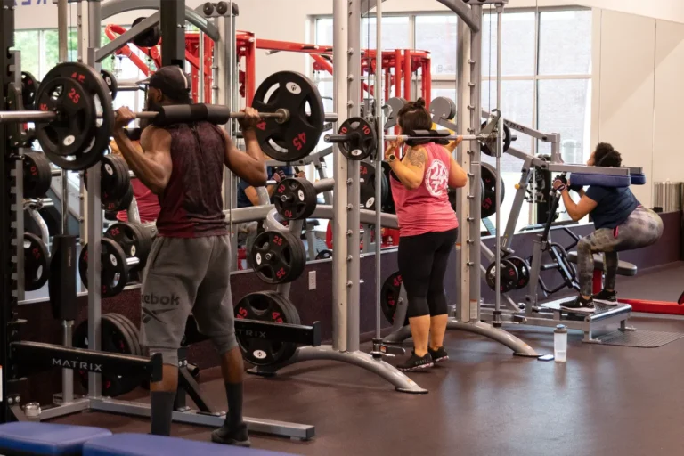 Three people at a gym – a man on the left and a woman in the middle do barbell squats, a woman on the right does the bodyweight squat.