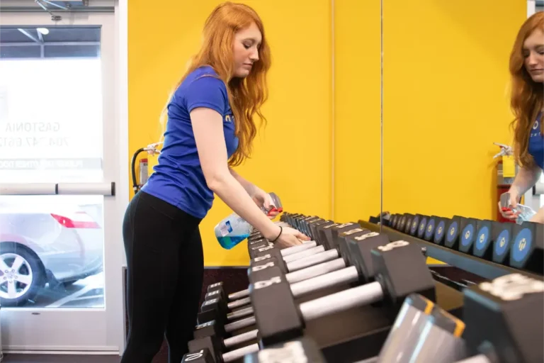 A red-haired woman in a blue t-shirt sanitizes weights with a spray bottle with blue liquid.