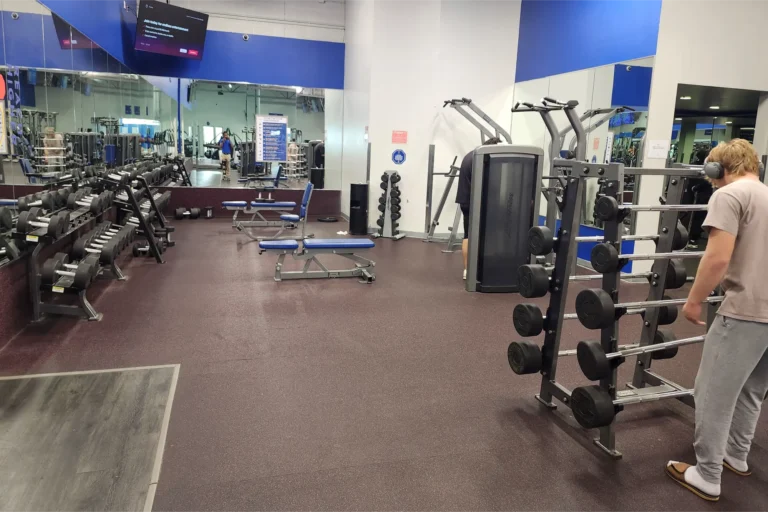 A man with headphones stands in front of a weight rack with more weight racks and benches in the background.