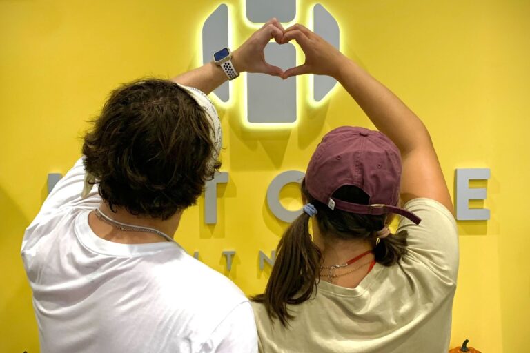 A man and a woman making a heart shape with their hands while their backs are seen. The woman wears a burgundy cap.