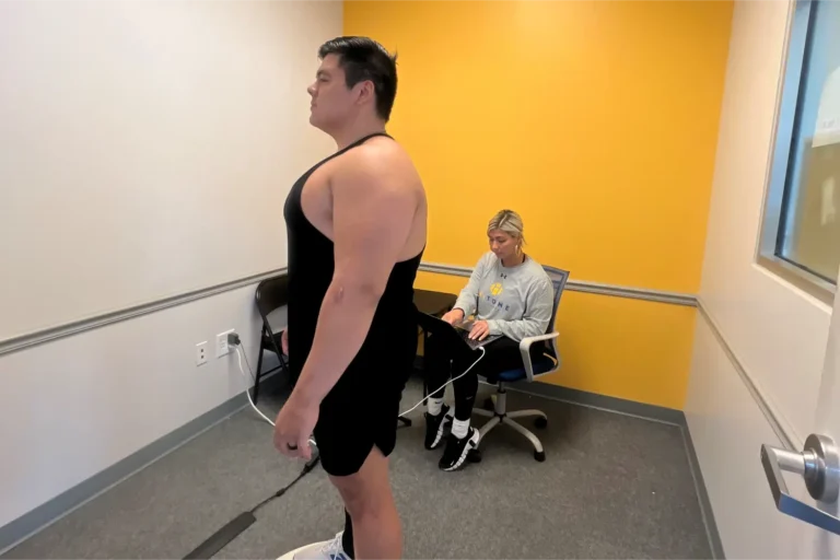 A man in black tank top stands near a woman working at a laptop from a chair.