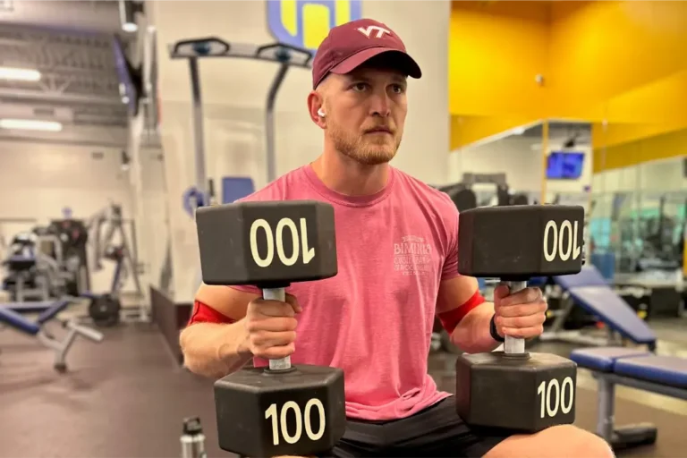 A man with a red cap and pink t-shirt is sitting at a gym and holding two 100-pound weights.