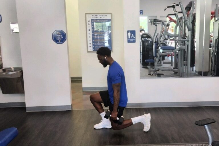 A man in a blue t-shirt with an earphone in his ear is doing a weighted lunge exercise at a gym.