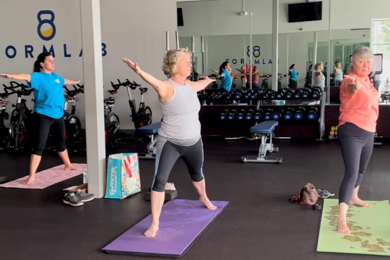 Two older women and a woman in her 30s doing a warrior II yoga pose at a gym.