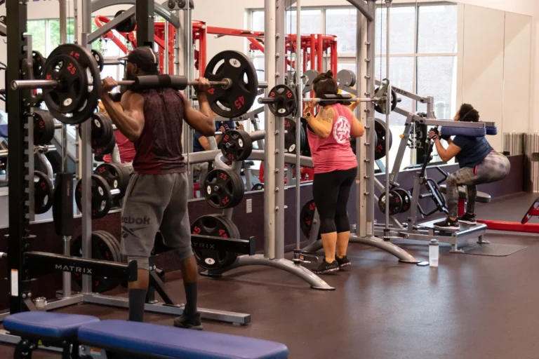 Three people at a fitness center – a man on the left and a woman in the middle do barbell squats, a woman on the right does the bodyweight squat.