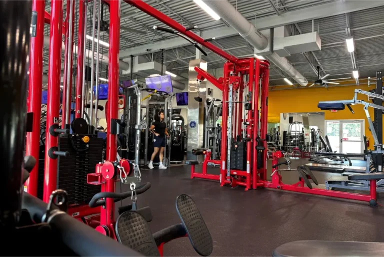 A red cable crossover machine at a fitness center. A woman is exercising on one in the background.