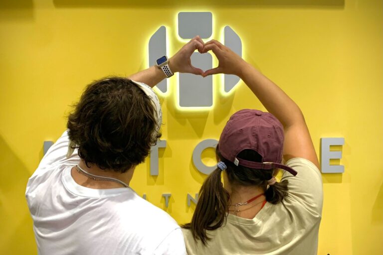 A man and a woman making a heart shape with their hands. Their backs are seen. The woman wears a burgundy cap.