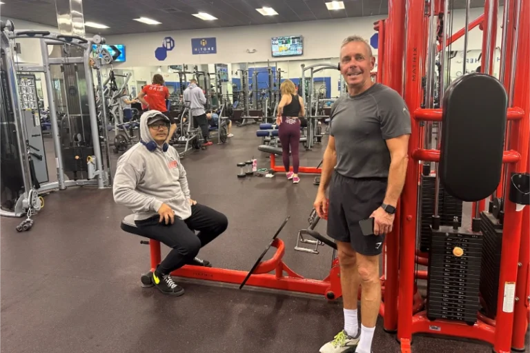 Two men at a gym looking at the camera. One is standing and one is sitting at a functional trainer machine.