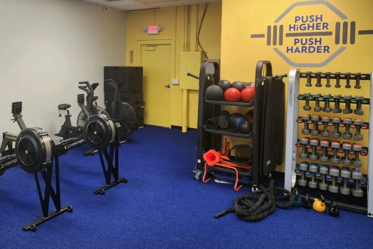 Rowing machines, and racks with medicine balls and dumbbells inside a room with yellow walls and blue carpet.