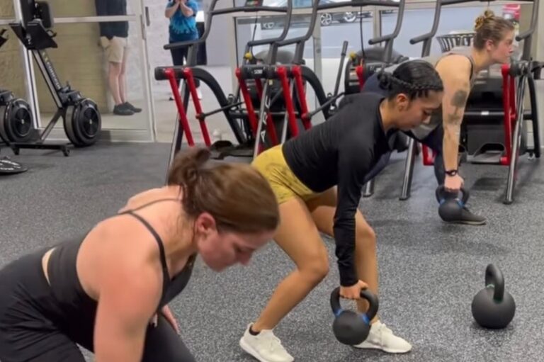 Three women doing the singlehanded kettlebell deadlift exercise inside a gym.