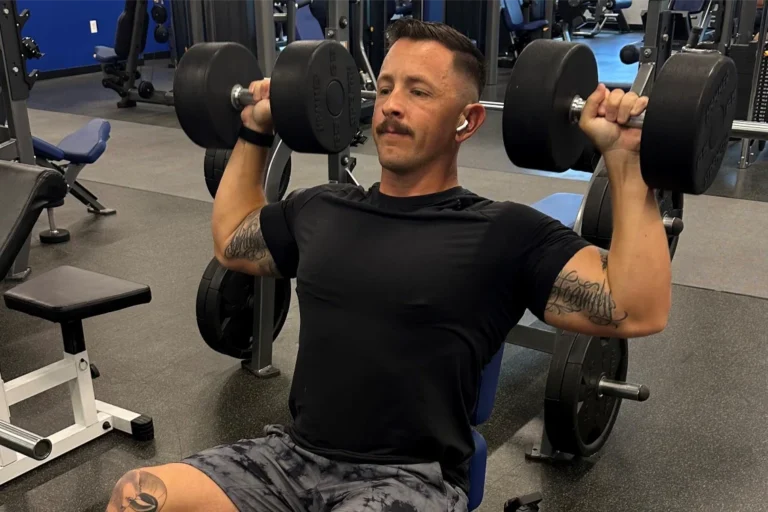 A man with a moustache doing the seated shoulder press using heavy dumbbells.