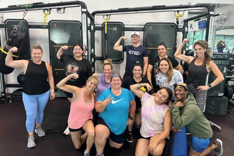 Eleven women and one man smiling and flexing their biceps in front of a functional training rig.