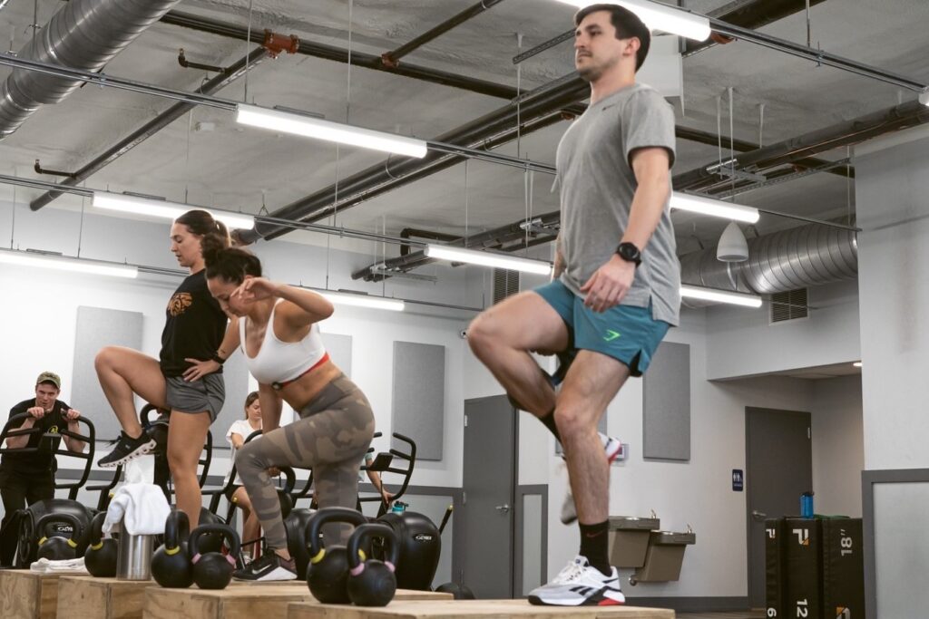 A man and two women doing step-ups on a box platform. A man and a woman in the background use the air bike.