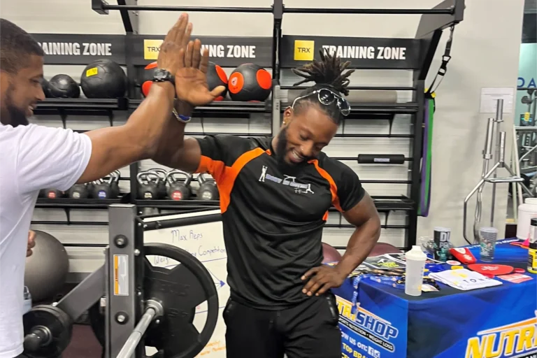 Two men doing high five at a gym. Rack with medicine balls and kettlebells is behind them.