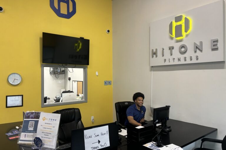 A man sitting at an office desk. HiTONE fitness wall sign is on his left above him.