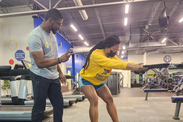 A woman doing the cable rope pull exercise while a fitness trainer counts her repetitions while standing behind her.