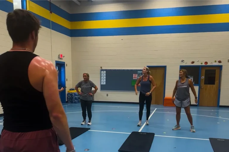 Three women standing in front of yoga mats. Man with a tank top stands in the foreground.