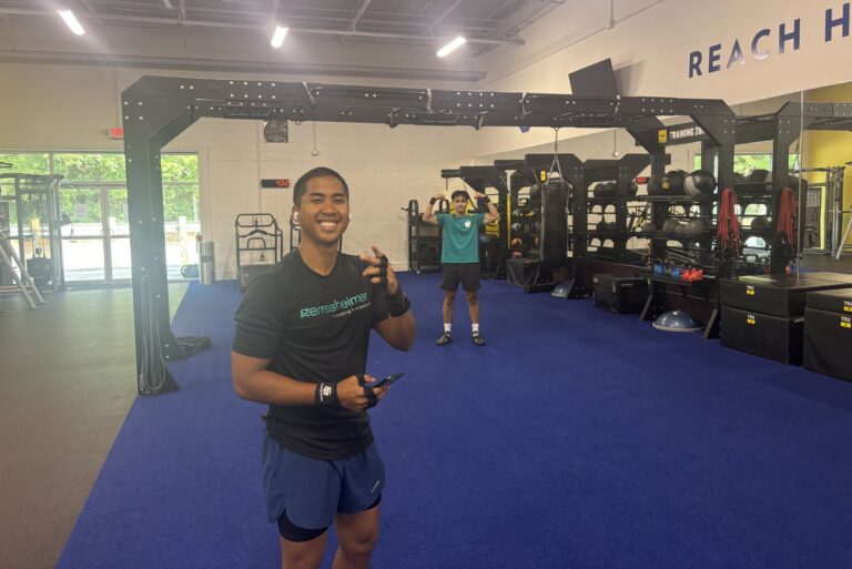 A smiling man pointing at a camera. Behind him, another man flexes arms under the functional training rig.