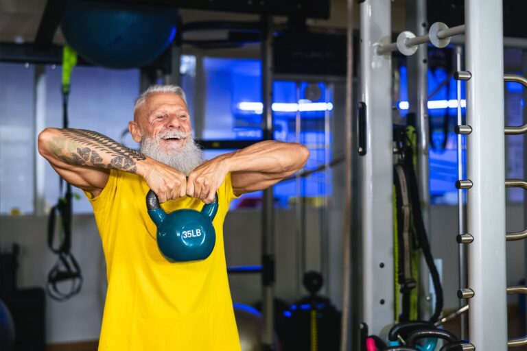 A bearded smiling man does the kettlebell upright row exercise at a gym.