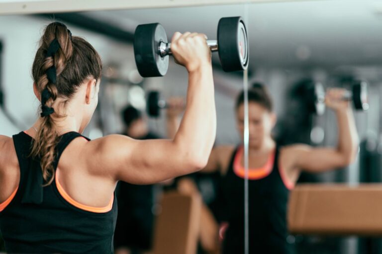 A woman doing the dumbbell shoulder press exercise in front of a window.