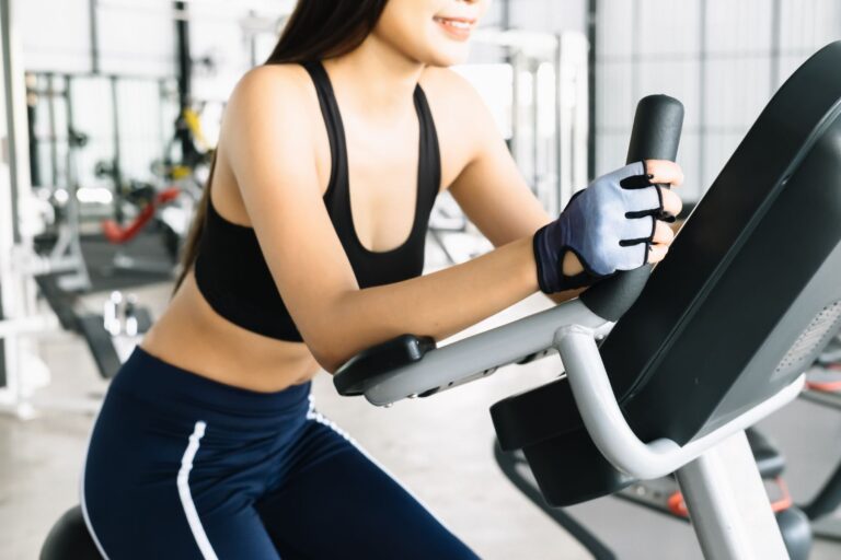 A smiling woman in a black cropped fitness tank top holds the handle of an exercise machine at a gym.