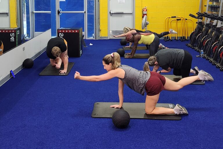 Four women and a man doing the bird dog yoga pose in a room with a mirror.