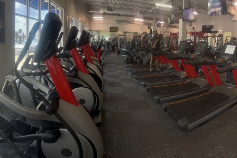 Elliptical machines and treadmills inside a gym with carpeted floor.