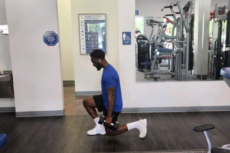 A man in a blue t-shirt with an earphone in his ear does the weighted lunge exercise at a gym.