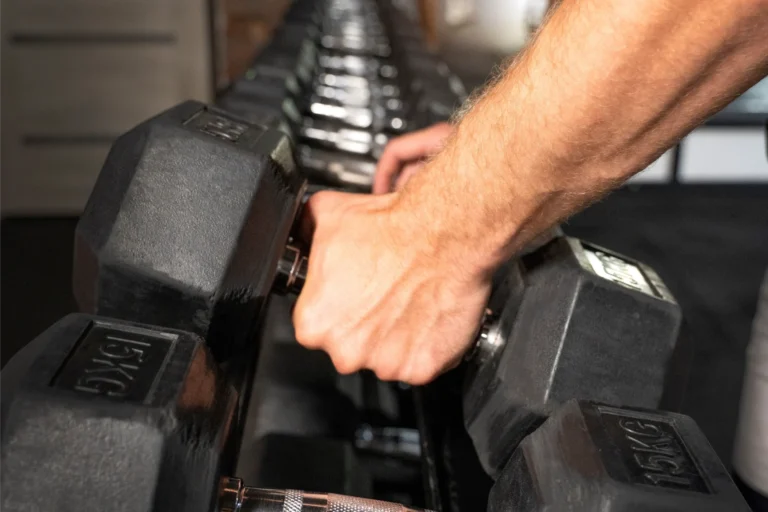 Men's hand gripping the handle of a hex dumbbell that stands on a rack.