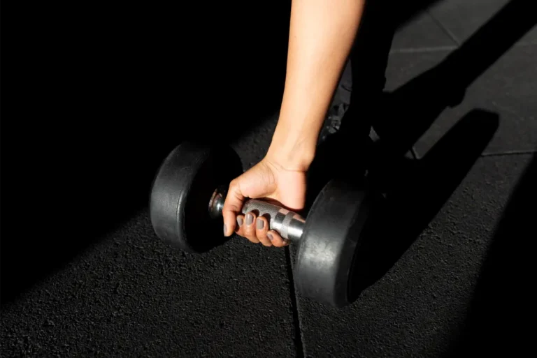 A woman's hand gripping the handle of a dumbbell placed on the concrete floor.