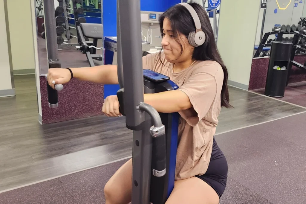 A woman with headphones doing the chest fly exercise on a pec deck machine.