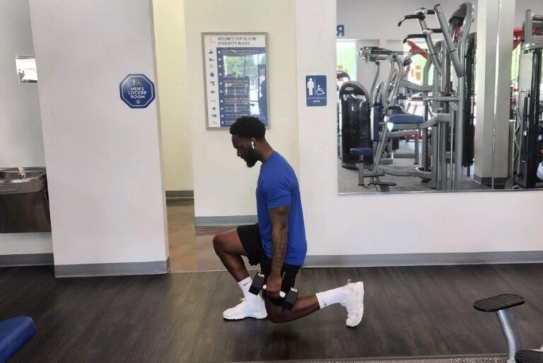 A man in a blue t-shirt with an earphone in his ear performs the weighted lunge exercise at a fitness center.