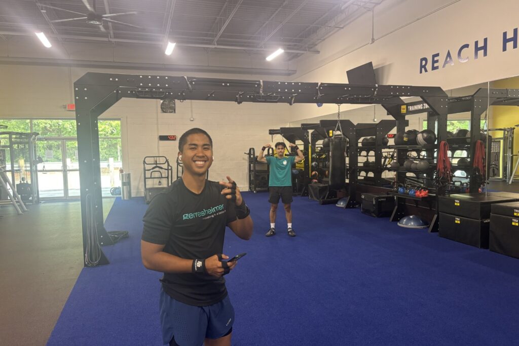 A man in the foreground in a black t-shirt is smiling while holding the phone. In the background, under the functional training station, a man is flexing biceps.