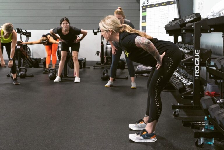 Five women doing stretching exercises next to racks with dumbbells and rowing machines.