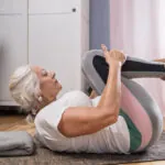 A senior woman in colorful yoga pants performs the knee-to-chest yoga pose on a mat inside a room.