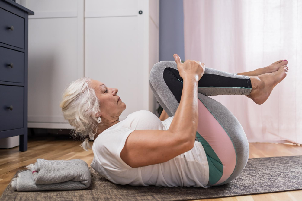 A senior woman in colorful yoga pants performs the knee-to-chest yoga pose on a mat inside a room.