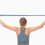 A woman in a gray workout tank holds a blue resistance band with both hands, extending it behind their head.