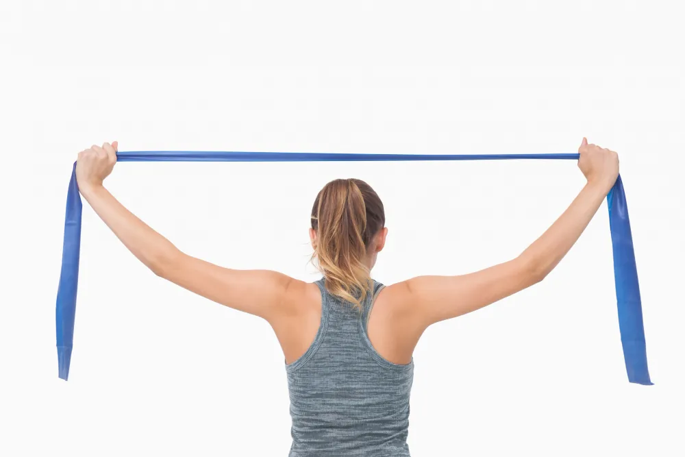 A woman in a gray workout tank holds a blue resistance band with both hands, extending it behind their head.