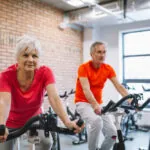 A senior woman and a man ride stationary bicycles at a gym. She wears a red t-shirt, he wears an orange t-shirt.