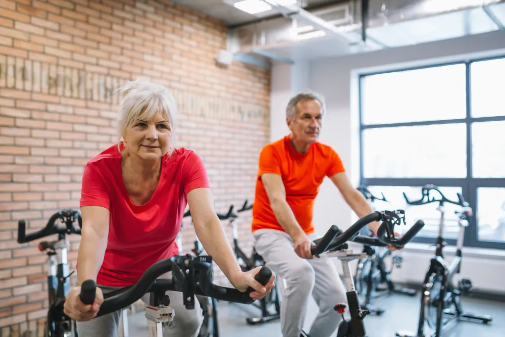 A senior woman and a man ride stationary bicycles at a gym. She wears a red t-shirt, he wears an orange t-shirt.