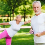 A man in a white t-shirt holds a clipboard while a woman in pink pants and a white shirt performs a stretching pose in a park.