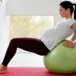 A pregnant woman in a gray shirt and maroon leggings rests on a green stability ball while exercising on a yoga mat.