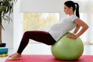 A pregnant woman in a gray shirt and maroon leggings rests on a green stability ball while exercising on a yoga mat.