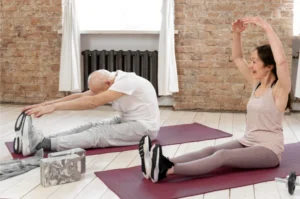 A senior man and a woman stretching on yoga mats in a well-lit room. A woman smiles with her arms up, a man touches his toes.