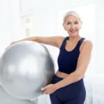 A senior woman in a navy tank top smiles while holding a silver exercise ball with one hand in a bright room.