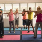 Six seniors (3 women and 3 men) are doing the upper body stretch while standing on yoga mats inside a daylight-lit room.